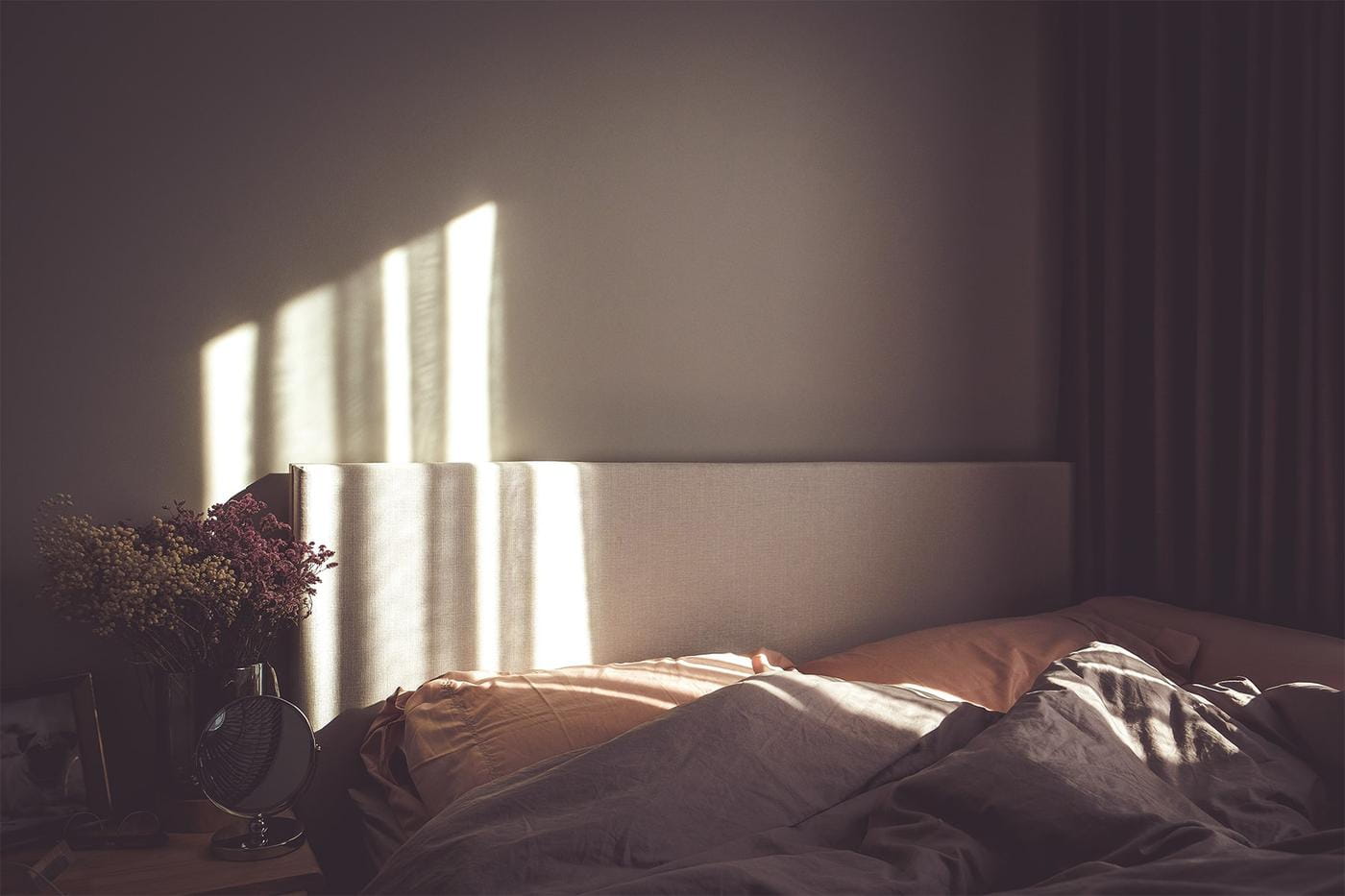 A disheveled bed in a dark room with the sun peeking through the window curtains, slightly illuminating the scene.